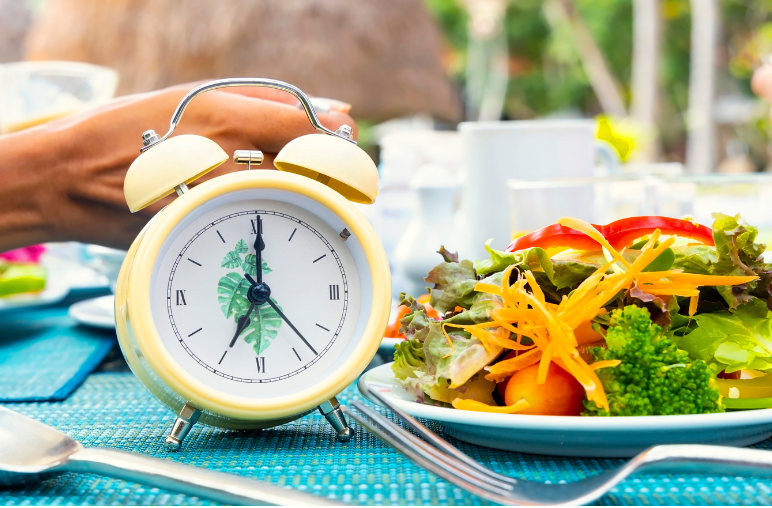 Yellow alarm clock next to a colorful salad, symbolizing the timing of fasting for autophagy.